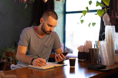 bearded cheerful male barista typing on phone while standing at bar counter in cafe. Job concept.の写真素材