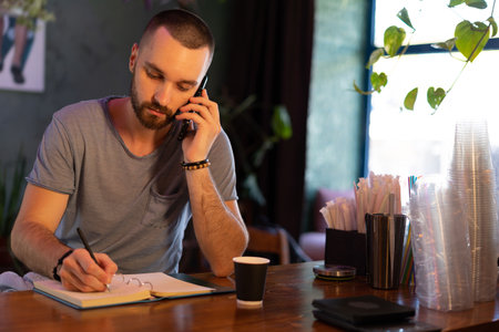 Confident bar owner. handsome young male bartender leaning at the bar counter writing something in note pad and have call on mobile phone while sitting at bar counterの写真素材