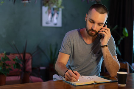 Confident bar owner. handsome young male bartender leaning at the bar counter writing something in note pad and have call on mobile phone while sitting at cafeの写真素材