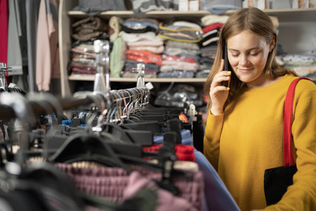 Smilling woman customer shopping in clothing store, using smartphone, choosing stylish clothes. Fashionable shop, colorful brands, sustainable designs.の写真素材