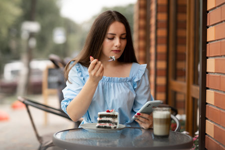 young woman eating cake at outdoors cafe and read message on her smart phone.の写真素材