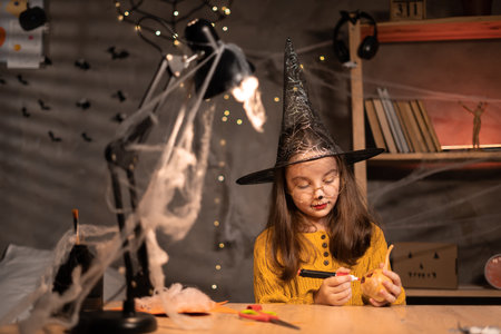 Cute young girl sitting at a table drawing a face on a small Halloween pumpkin. Halloween holiday and family lifestyle background. Holiday decorationの写真素材