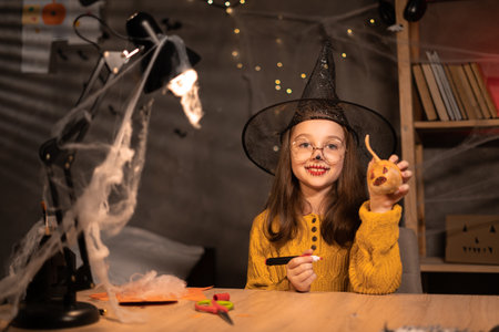 Smilling little girl preparing for halloween drawing scary face on pumpkin while sitting at table at home, smiling child making jack-o-lantern. Holiday decorationの写真素材