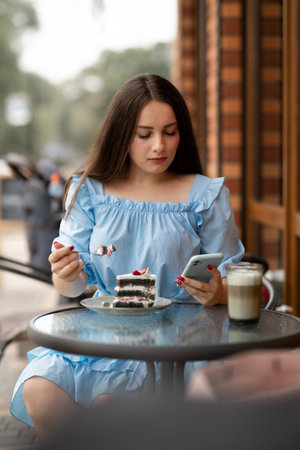 Beautiful woman eating cake at outdoors cafe and read message on her smart phone.の写真素材