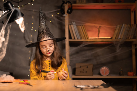 Cute happy little witch preparing for Halloween, painting drawing scary face on pumpkin while sitting at table at home, smiling child making jack-o-lantern. Holiday decorationの写真素材