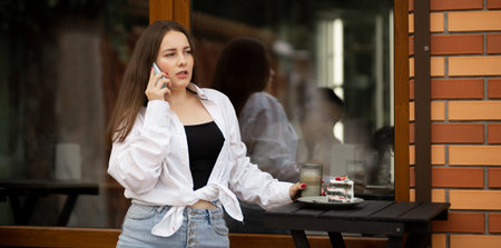 Thoughtful caucasian woman calling on mobile phone while looking near the coffee shop window during coffee break. Street reflections in the window glass. woman talking on mobile phone outdoors.の写真素材