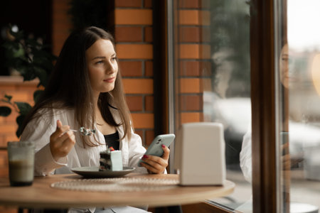 Young woman having breakfast in a cafe and using a mobile phone for chatting, looking thoughtfully out the window, happy leisure time in a city cafeの写真素材