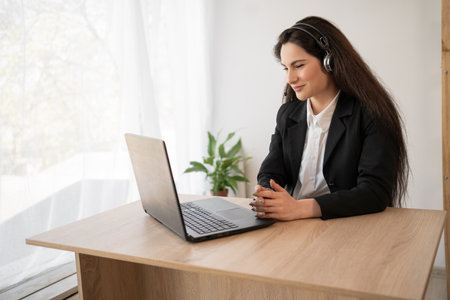 Latin woman in headphones working online using laptop in office, serious millennial female employee in headset consult client on Internet on computer, watching films online on her pcの写真素材