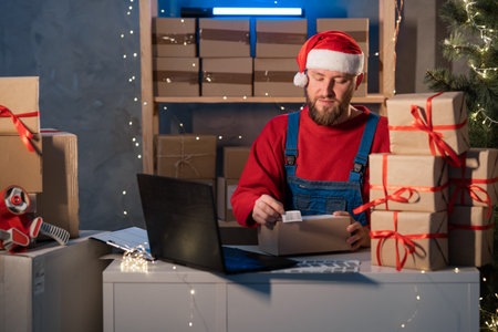 Caucasian business owner working with product packaging and laptop wearing a Sanat Claus hat and red sweater. Glue barcode on box and prepares purchase for shipment. Christmas saleの写真素材