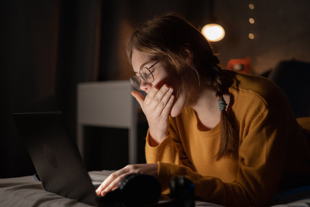 Tired young woman yawning, lying on bed with laptop, lazy student doing homework, sleepy girl working on computer in dormitory, sleepless night, lack of sleep and boredom conceptの写真素材