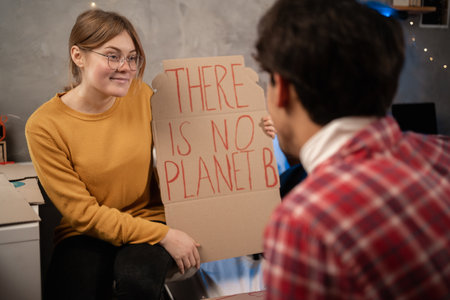 Students prepare banner protesting over pollution and global warming sitting in dormitory. Protest to save planet earth. Woman showing a banner of There is no Planet B for her boyfriend. close upの写真素材