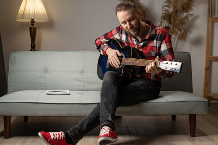 Bearded young man playing the guitar while sitting on sofa at home in the evening. copy spaceの写真素材