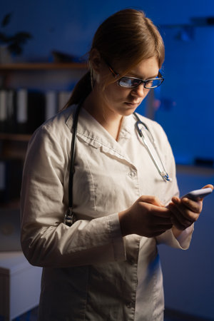 Shot of pretty female doctor using her mobile phone while standing in medical consultation during night shift. copy spaceの写真素材