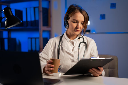 Young tired doctor working night shift in office with papers, therapist with headset holding video call with patient. close upの写真素材