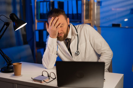 Male tired doctor with migraine overworked, overstressed working at night. sad unhappy health care professional with headache stressed sleepy. copy spaceの写真素材