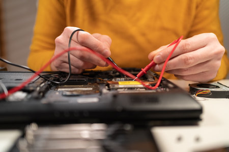 Close-up of man hands testing and examining laptop motherboard with multimeter at desk. Maintenance, repairing and checking computer hardware concept.の写真素材
