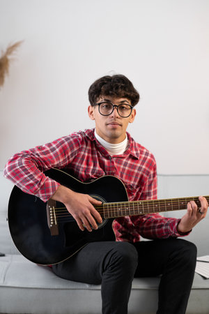 Handsome young man in casual clothes is looking at camera while playing guitar at home. copy spaceの写真素材
