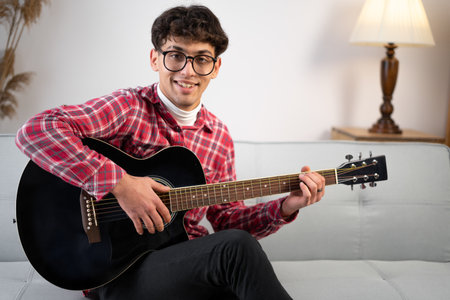 Attractive relaxed man sitting on a couch and playing the guitar looking at camera. He is smiling at the camera. horizontal shot. copy spaceの写真素材