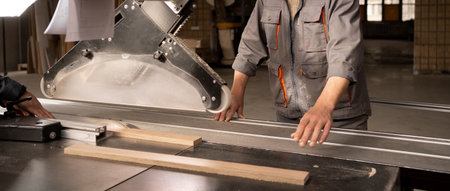 Male craftsman saws wood on a circular saw, carpenter saws wood on a saw machine in furniture factory. Production, manufacture and woodworking industry concept. copyspace. Bannerの写真素材
