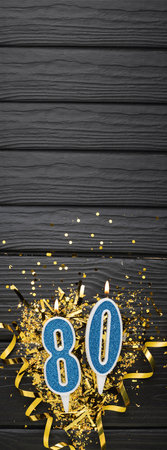 Number 80 blue celebration candle and gold confetti on dark wooden background. 80th birthday card. Anniversary and birthday concept. vertical banner. copy spaceの写真素材