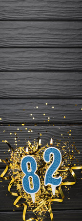 Number 82 blue celebration candle and gold confetti on dark wooden background. 82th birthday card. Anniversary and birthday concept. vertical banner. copy spaceの写真素材