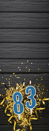 Number 83 blue celebration candle and gold confetti on dark wooden background. 83th birthday card. Anniversary and birthday concept. vertical banner. copy spaceの写真素材
