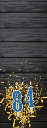 Number 84 blue celebration candle and gold confetti on dark wooden background. 84th birthday card. Anniversary and birthday concept. vertical banner. copy spaceの写真素材