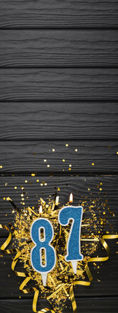 Number 87 blue celebration candle and gold confetti on dark wooden background. 87th birthday card. Anniversary and birthday concept. vertical banner. copy spaceの写真素材