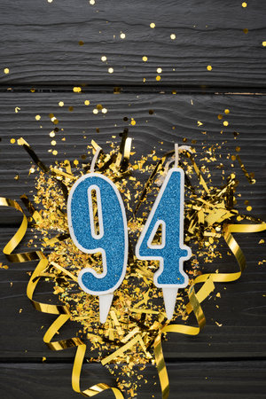 Number 94 blue celebration candle and gold confetti on dark wooden background. 94th birthday card. Anniversary and birthday concept. vertical banner. copy spaceの写真素材
