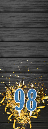 Number 98 blue celebration candle and gold confetti on dark wooden background. 98th birthday card. Anniversary and birthday concept. vertical banner. copy spaceの写真素材