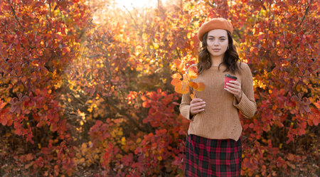 Beautiful young happy girl with a bright red-yellow autumn leaf and cup of coffee in the parkの写真素材