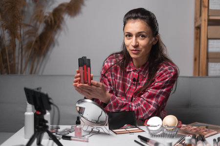 Beauty blogger woman filming on smartphone daily make-up routine tutorial. Influencer girl live streaming cosmetics product review in home studio. Vlogger female comparing lipsticks.の写真素材