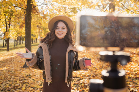 Female blogger recording video outdoors. Beautiful young girl smiling and looking at the camera standing in the autumn park, talking and gesticulating with both hands. Blogging, video blogの写真素材