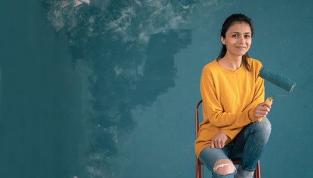 Smiling nice girl, in an orange sweater, holding a paint roller in her left hand and looking at the camera. Time to paint.の写真素材