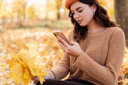Portrait of smiling woman using mobile phone in park during the autumn, sitting on a ground with orange leaves.の写真素材