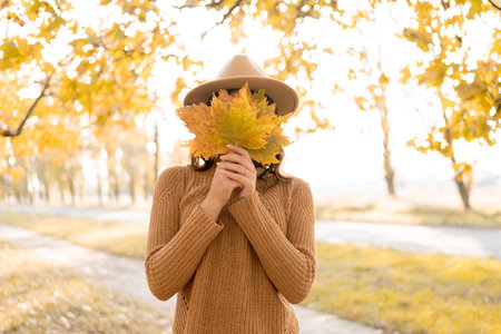 Beautiful happy young woman in park in fall holding leaves close her face. Cheerful girl outdoors in autumn.の写真素材