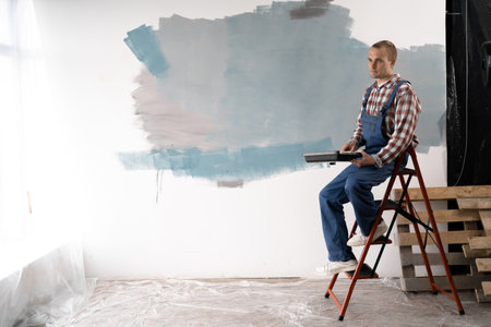 Young worker painting wall in room, sitting on ladder resting after work. Painter doing repair in homeの写真素材