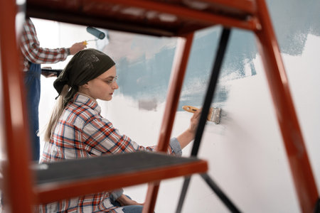 Profile view of young family married couple doing renovating works in house. Wife and husband painting walls together redecorating and redesigning homeの写真素材