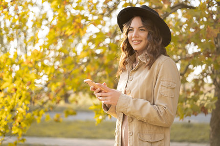 Autumn portrait smiling woman holding phone over yellow maple leaves background. Happy girl using a...