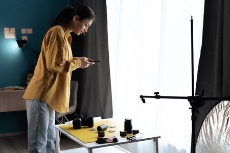 Female product photographer with smartphone and camera photographing cosmetic products in home photo studio. Professional and mobile photo conceptの写真素材