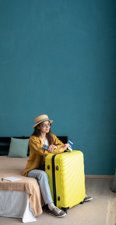 Smiling young woman in casual clothes and hat relaxing with a yellow suitcase in her bedroom. Time to go to travel in a summer time. Banner. copy spaceの写真素材
