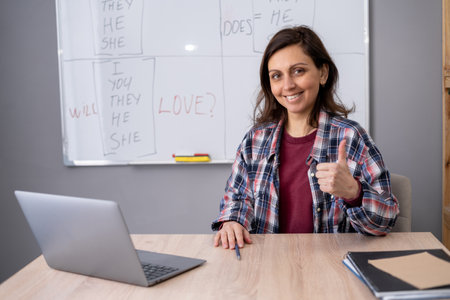 Female teacher and student having online conference, using laptop computer. Woman tutor foreign lesson smiling and showing thumb up satisfying with class results sitting at deskの写真素材