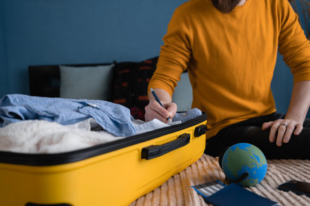 A man in a yellow sweater writes down what to take with him on a trip. Making check list of things to pack for travel, prepare clothes into luggage.の写真素材