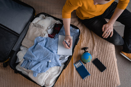 Making a check list of things to pack for travel. Top view of man writing paper take note and packing suitcase to vacation, prepare clothes into luggage. Travel vacationの写真素材