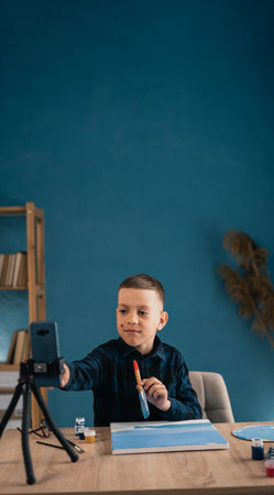 Caucasian guy schoolboy at home online learns to draw with a brush and paints on canvas looks at the phone remotely video lesson. Childrens education, entertainment, hobby, creativity concept.の写真素材