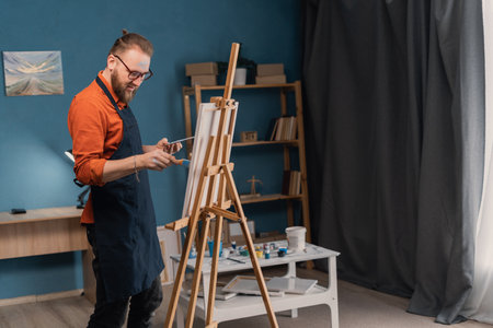 Bearded male artist working on painting on canvas on the easel at home interior. Man artist painter in creative studio as art conceptの写真素材