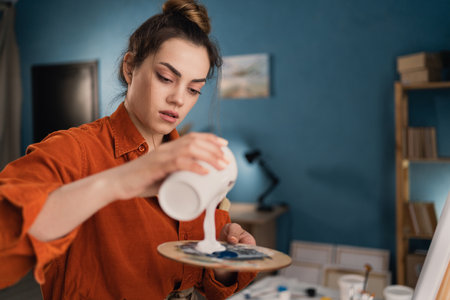 close-up portrait of a young brazilian woman artist in a home studio standing near a canvas pouring white paint on a palette. Black hair, dressed in casual clothes. concept of creativity and hobbyの写真素材