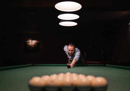 A bearded caucasian man stands near the billiard table, prepares for the start of the game, bowed down, holds a cue in his hands, takes aim. Billiard sport concept.の写真素材