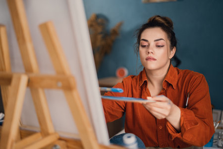 a concentrated Brazilian woman artist with paint on her hands works on drawing a picture on canvas sitting in front of her, applying a brushstroke on a stool. home studio. hobby and vacation conceptの写真素材