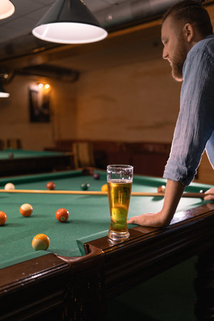 Serious man standing near pool table with fresh beer. A glass of cold beer and billiard balls on a...
